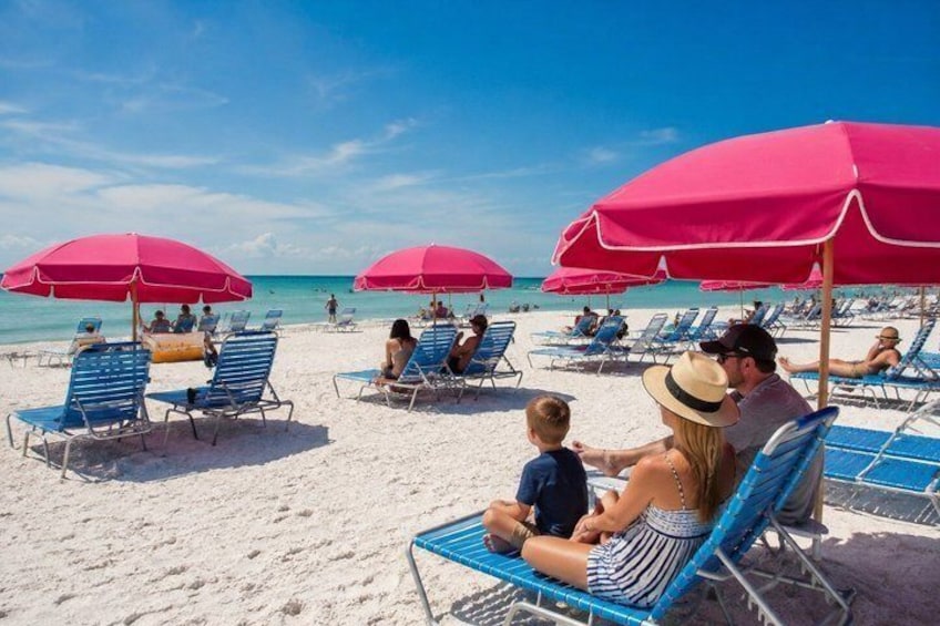 A beach with people relaxing under pink umbrellas, sun loungers, and a clear blue sky. 5