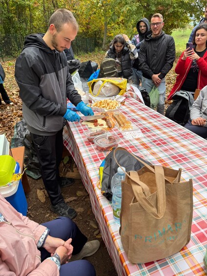A group of people gathered around a table with a checkered tablecloth, preparing food outdoors. 2