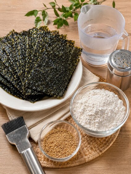Dried seaweed, a bowl of flour, a container of brown rice, a salt shaker, a water pitcher, and a strainer on a wooden surface. 1