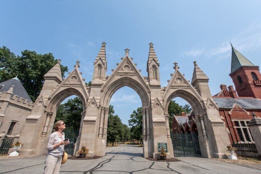 A Gothic-style archway with intricate stonework, a red brick building with a green roof, and a clear blue sky. 1
