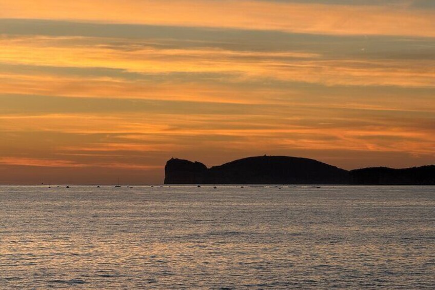 A sunset over a calm sea with a silhouette of a large rock formation and several boats. 2