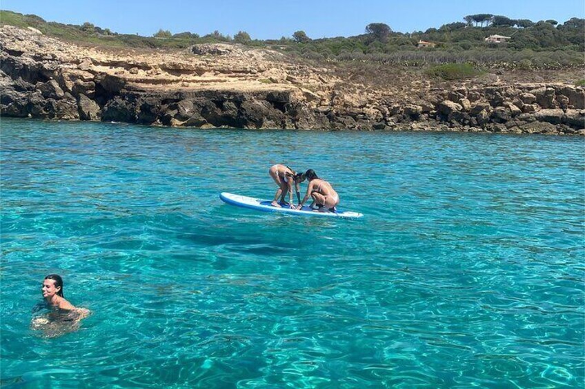 Two people on a paddleboard in clear blue water, with a rocky coastline and sparse vegetation in the background. 3