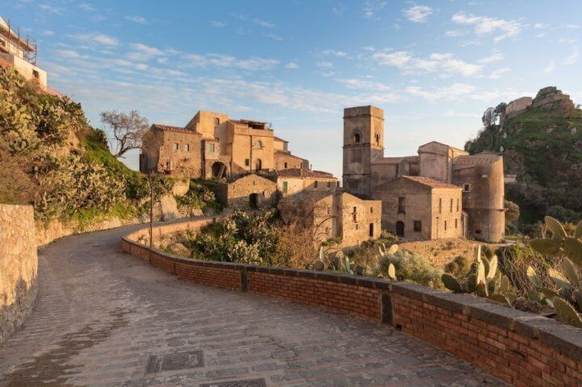 A historic stone pathway leading to a fortified building complex with multiple towers and walls, surrounded by cacti and shrubs. 2