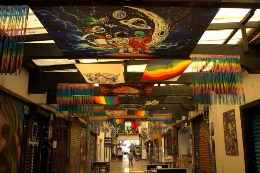 A narrow alleyway with colorful drapes hanging from the ceiling, a mural depicting space exploration, and closed shops on either side. 4