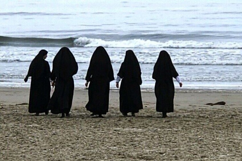 Four individuals in black robes walking on a beach with waves in the background. 4