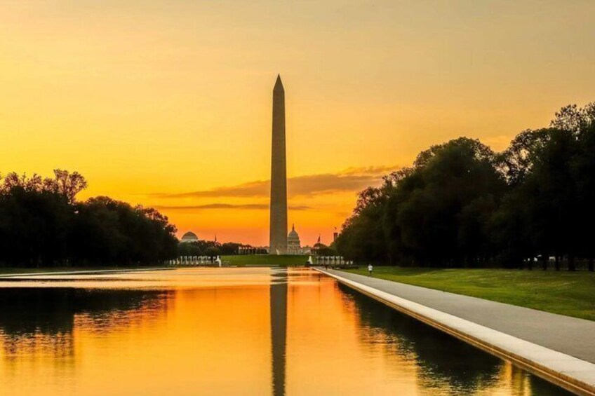 The Washington Monument at sunset with a reflection in the water, surrounded by trees and a pathway. 1