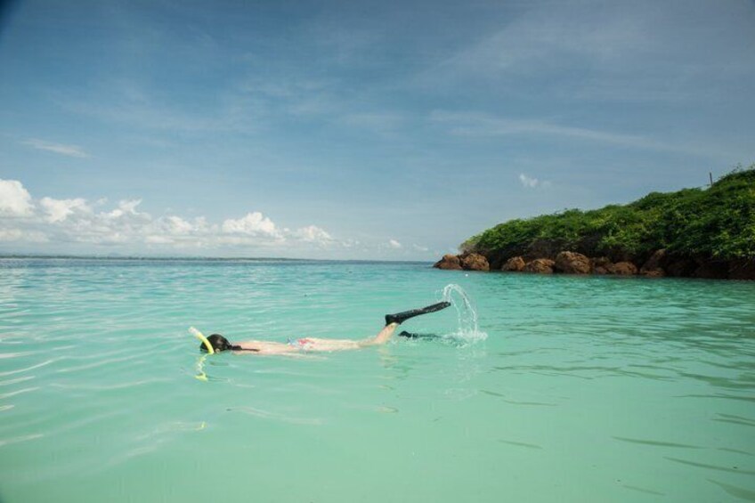 A person floating on a surfboard in clear turquoise water with a rocky coastline and greenery in the background. 2