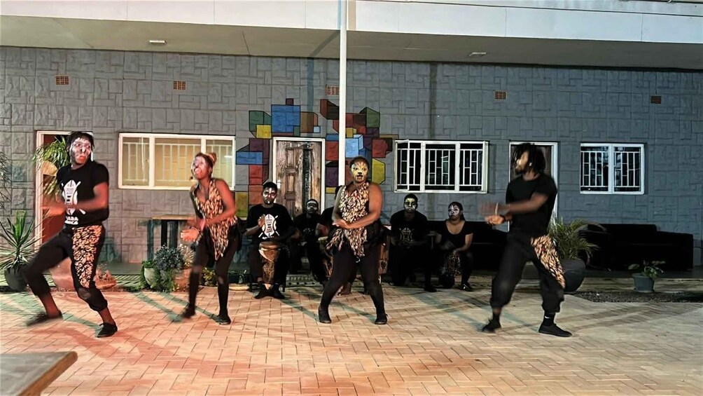 A group of people performing a dance in an outdoor setting with a brick building and windows in the background. 2