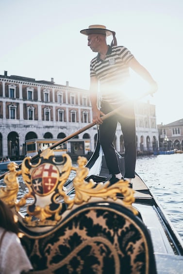 A gondolier in a striped shirt and straw hat standing on a gondola with ornate decorations, in a canal with historic buildings in the background. 3