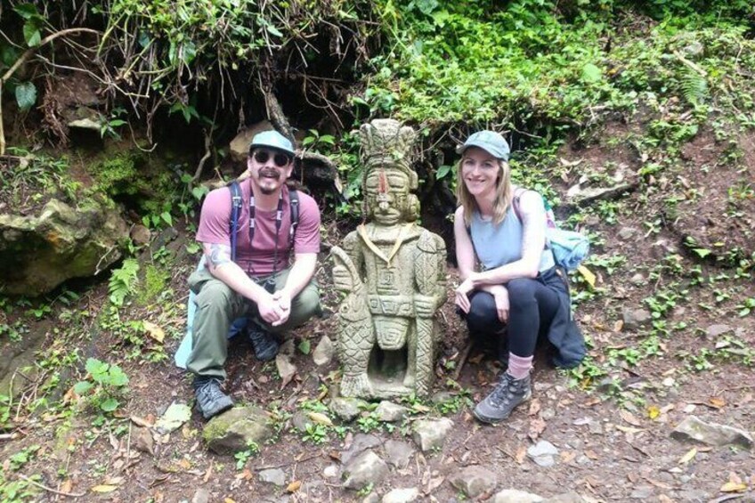 Two people sitting next to a stone statue in a forested area. 3
