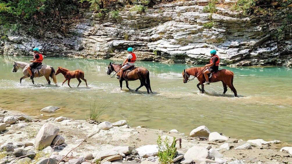 Three people riding horses through a shallow river surrounded by rocky terrain. 2