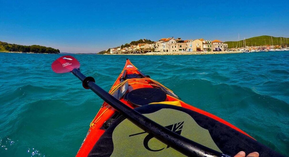 A kayak on turquoise water with a paddle, a red hat, and a green life vest. In the background, there is a coastal town with buildings and a hill. 3
