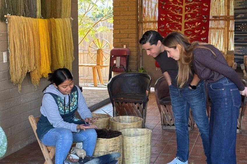 A person weaving with yarn on a chair, with baskets and a spinning wheel in the background. 2