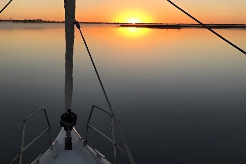 A boat's bow with a rope and a winch, a calm body of water, and a sunset in the background. 1