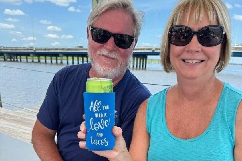 Two people holding a blue tumbler with text, standing by a waterfront with a pier in the background. 5