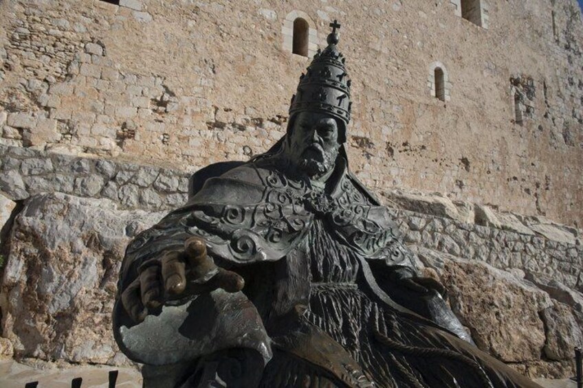 A statue of a robed figure with a pointed hat, set against a stone wall with arched windows. 5
