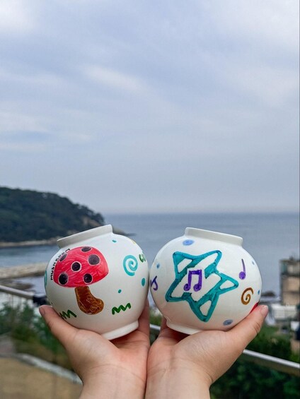 Two hand-held ceramic mugs with colorful designs, held against a backdrop of a coastal landscape with a body of water and a hilly terrain. 2