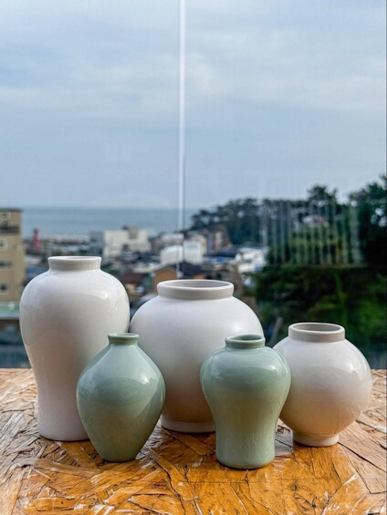 Five ceramic vases on a wooden surface with a coastal cityscape in the background. 5