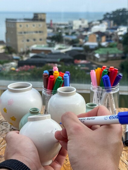 A person holding a ceramic marker, with colorful markers in jars and ceramic vases in the foreground, and a cityscape in the background. 3