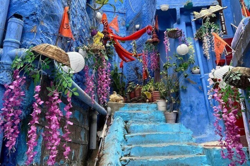 A narrow alley with blue walls, pink flowers, and orange flags. 1