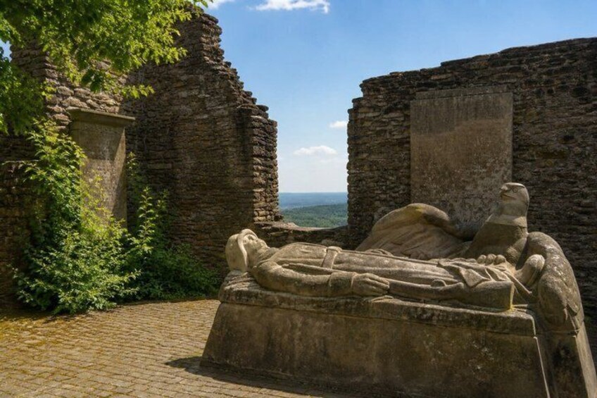 A stone sarcophagus with a reclining figure, set against a backdrop of ancient ruins and a clear sky. 2