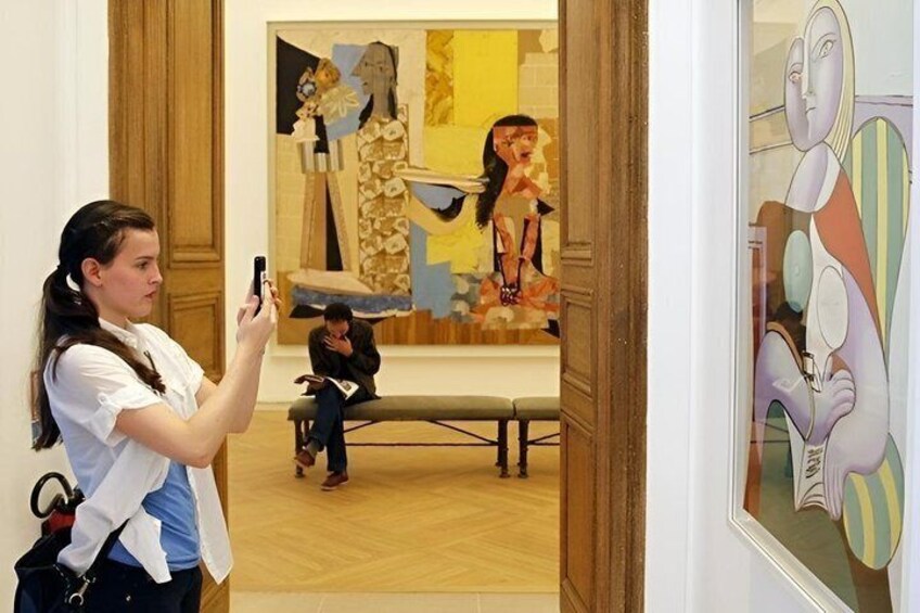 A person taking a photo with a smartphone, a painting with abstract figures, and a wooden floor. 1