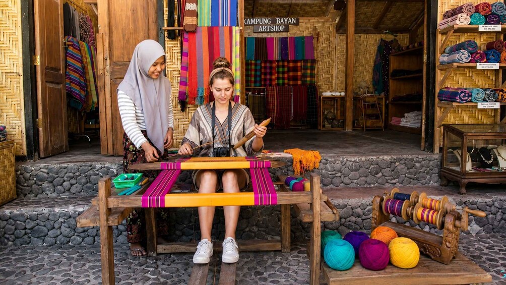 Two people weaving on a wooden bench outside a shop with colorful textiles and yarns displayed. 3