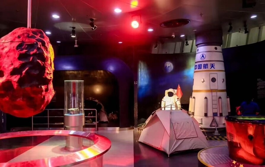A space-themed exhibit with a large red rock, a glass enclosure, a white rocket, and a tent. 5