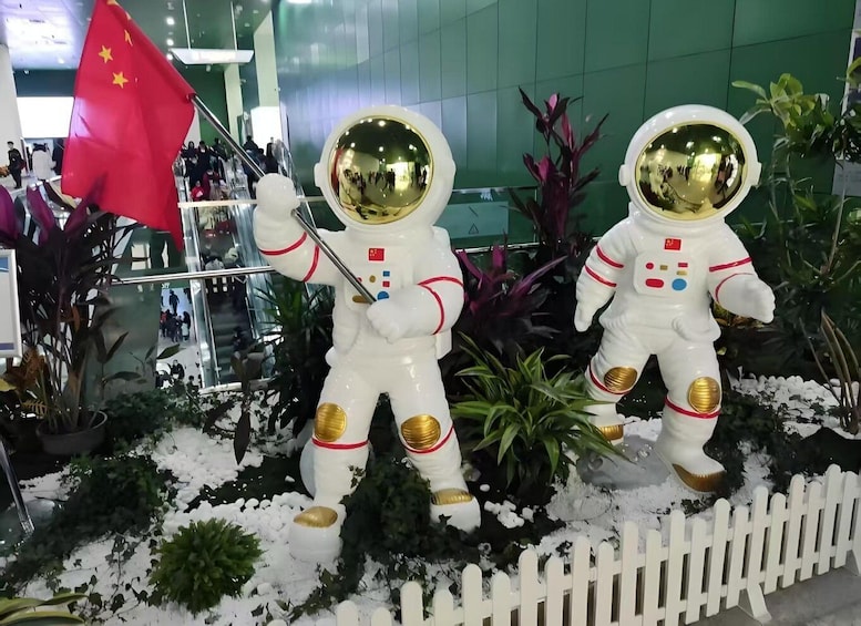 Two astronaut statues holding a flag and a pole, surrounded by plants and snow. 1
