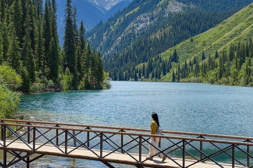 A person walking on a wooden bridge over a lake surrounded by dense forest and mountains. 1