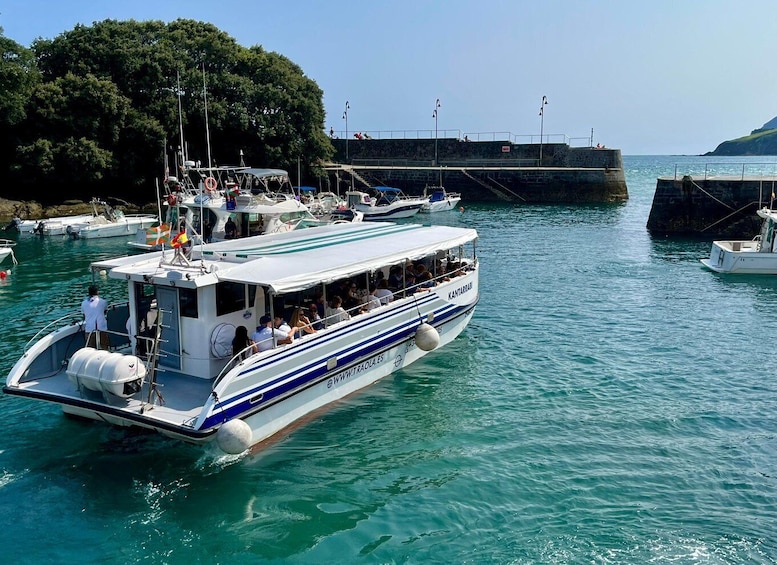 A boat with passengers docked at a marina with other boats and a rocky shoreline in the background. 1