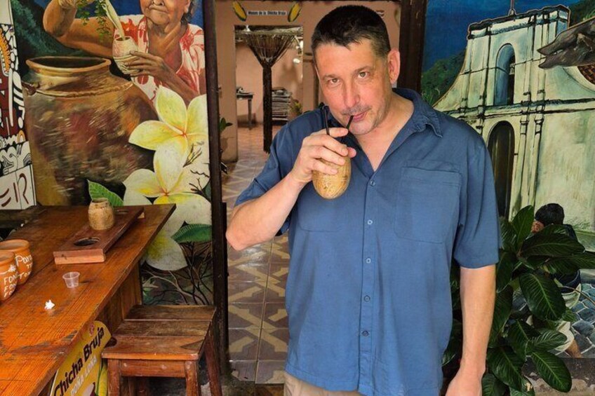 A man in a blue shirt holding a drink, standing in a room with a mural and a wooden table with a sign. 4