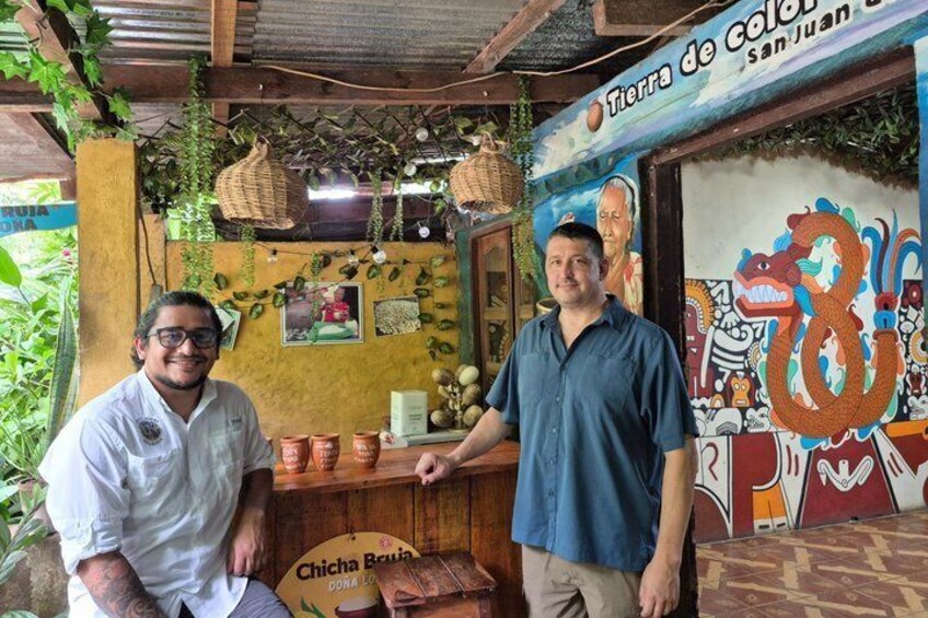 Two men standing in front of a rustic shop with a colorful mural, terracotta pots, and hanging baskets. 5