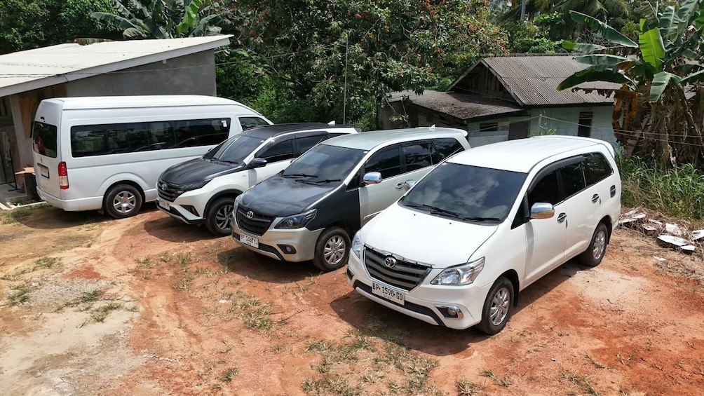 A white minivan parked on a dirt road with a bus and two cars in the background. 1