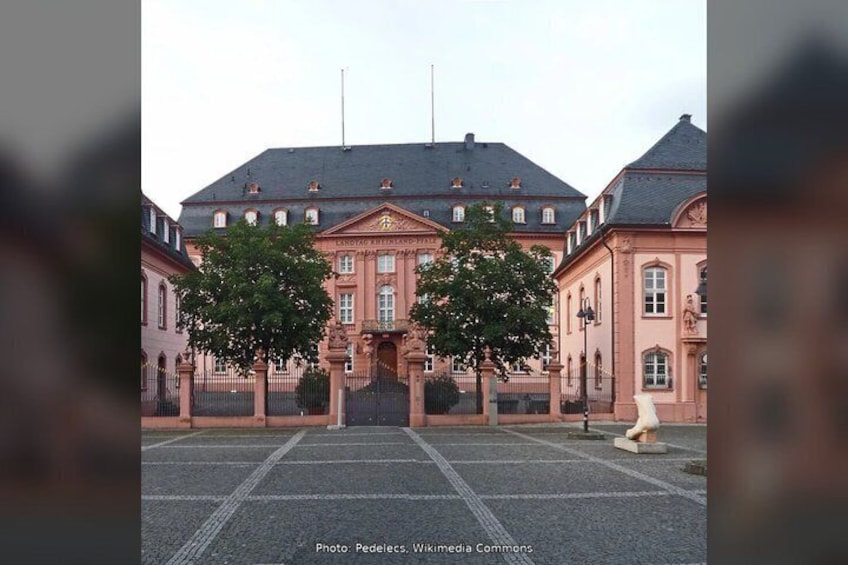 A large, historic building with a symmetrical facade, a central entrance, and two trees in the foreground. 2
