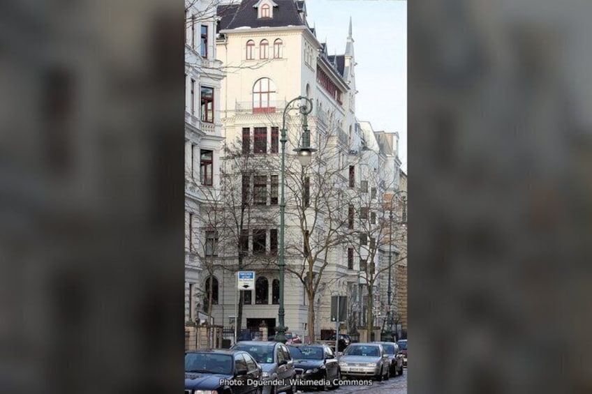 A street with parked cars, a building with multiple windows and a balcony, and bare trees. 3