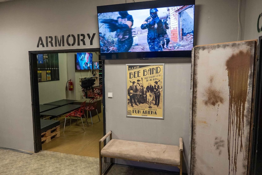 A room with a television mounted on the wall displaying a video game, a poster for a band performance, and a sign that reads 'ARMORY'. 4