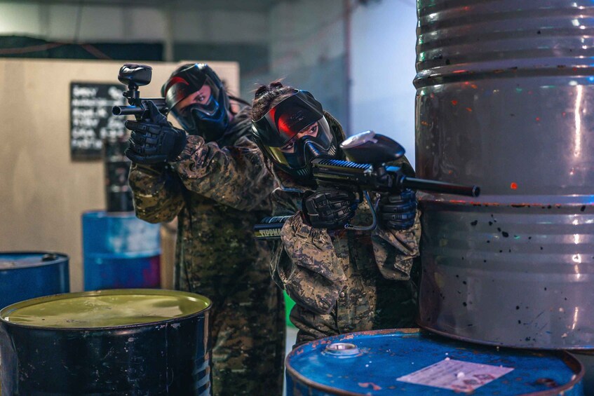 Two people in camouflage gear with paintball masks and gear, aiming paintball guns at a target. 3