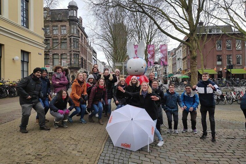 A group of people posing with a large white umbrella and a rabbit mascot in a cobblestone street with historic buildings. 2