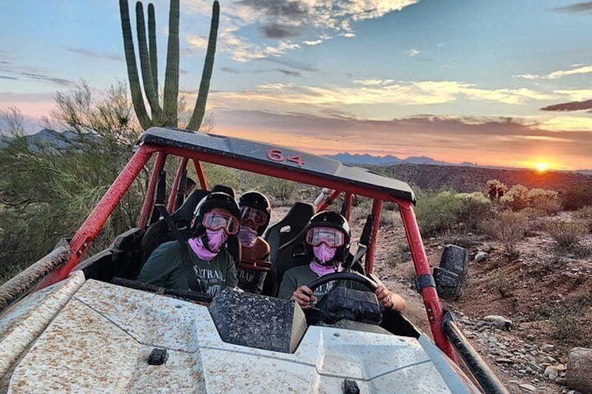 A group of people in a red off-road vehicle on a desert landscape with cacti and mountains in the background during sunset. 2