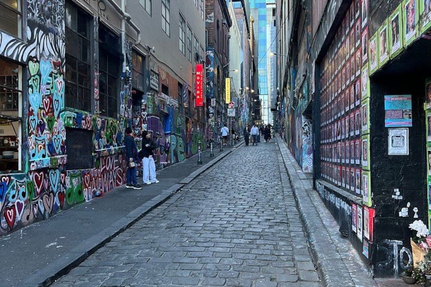 A cobblestone street with graffiti, a row of shops, and pedestrians. 1