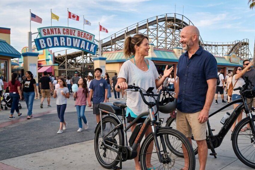 A crowded boardwalk with bicycles, a sign for Santa Cruz Beach Boardwalk, and a roller coaster in the background. 3