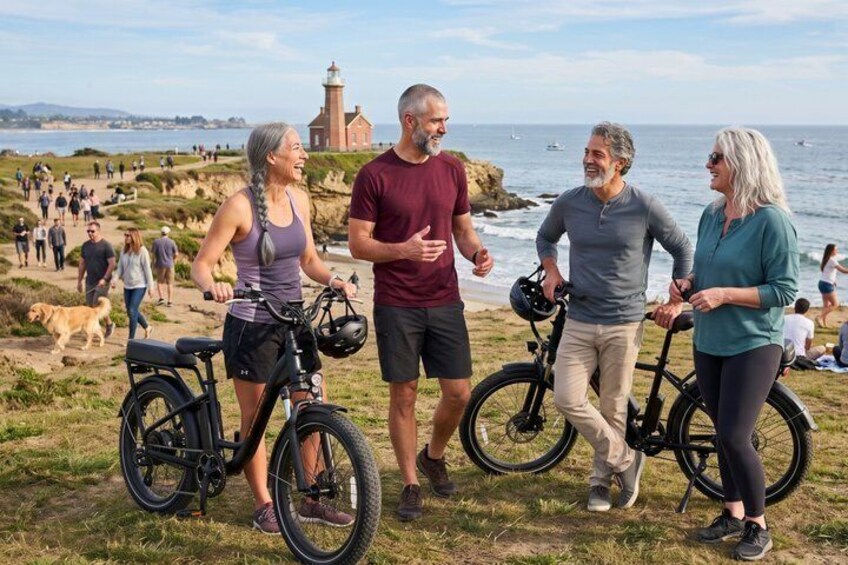 A group of people on a grassy area near the ocean with bicycles, a lighthouse in the background, and a crowd of people in the distance. 1