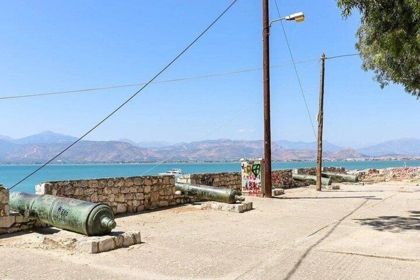 A coastal area with stone walls, a paved walkway, and large green cannons. 3