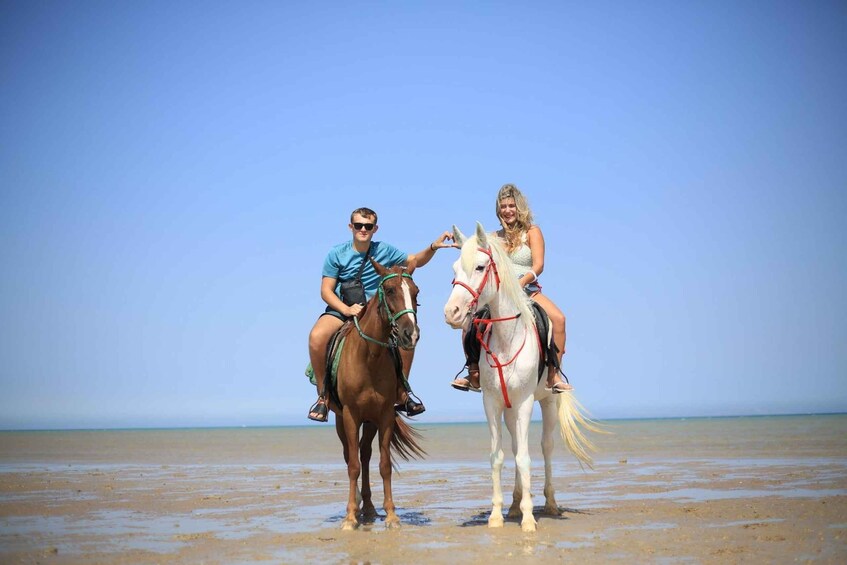 Two people riding horses on a beach with a clear sky. 2