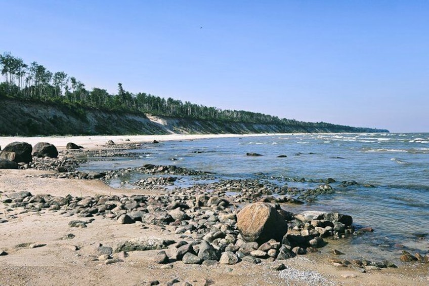 A rocky shoreline with clear water, a sandy beach, and a forested hill in the background. 1
