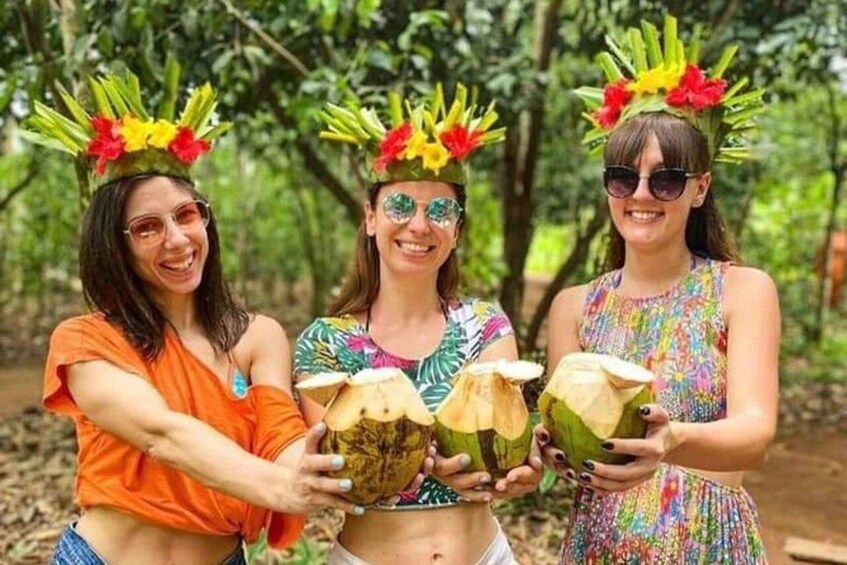 Three people holding coconuts with colorful headdresses in a forest. 5
