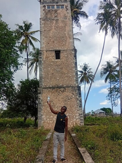 Una persona de pie frente a una torre alta y erosionada, con una pequeña ventana, rodeada de palmeras y follaje. 2