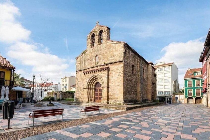 A historic stone building with a bell tower, a paved square with benches, and colorful buildings in the background. 3