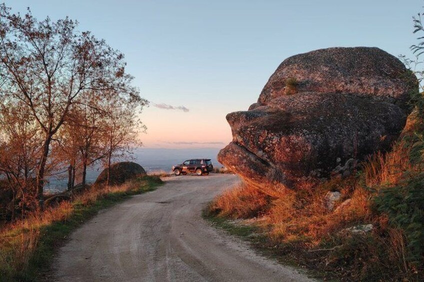 A dirt road with a large rock on the right and a vehicle in the distance. 1